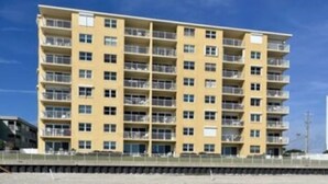 Un hotel de varios pisos con balcones, techo plano y terreno arenoso en la parte frontal.