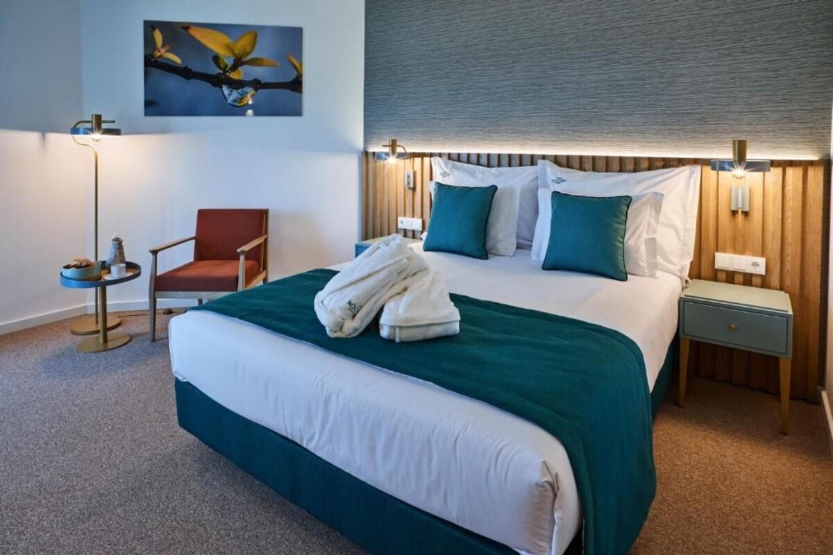 A hotel room with a large bed, a chair, a bedside table, a lamp, and a wall-mounted artwork.