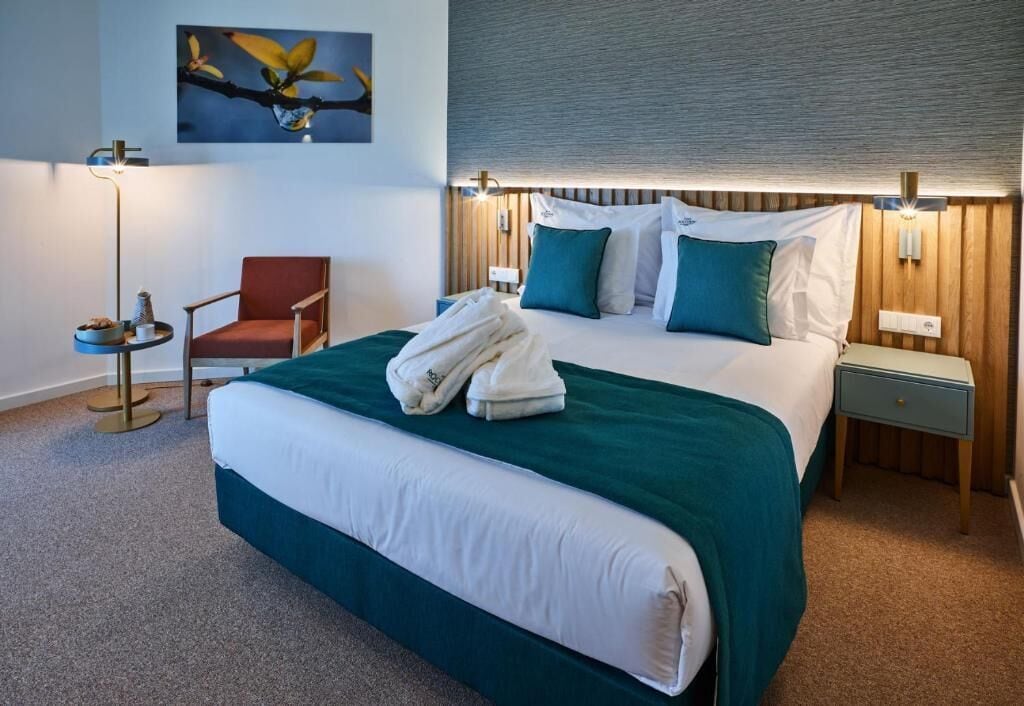 Ein Hotelzimmer mit einem großen Bett, einem Sessel, einem Nachttisch, einer Lampe und einem an der Wand hängenden Kunstwerk.