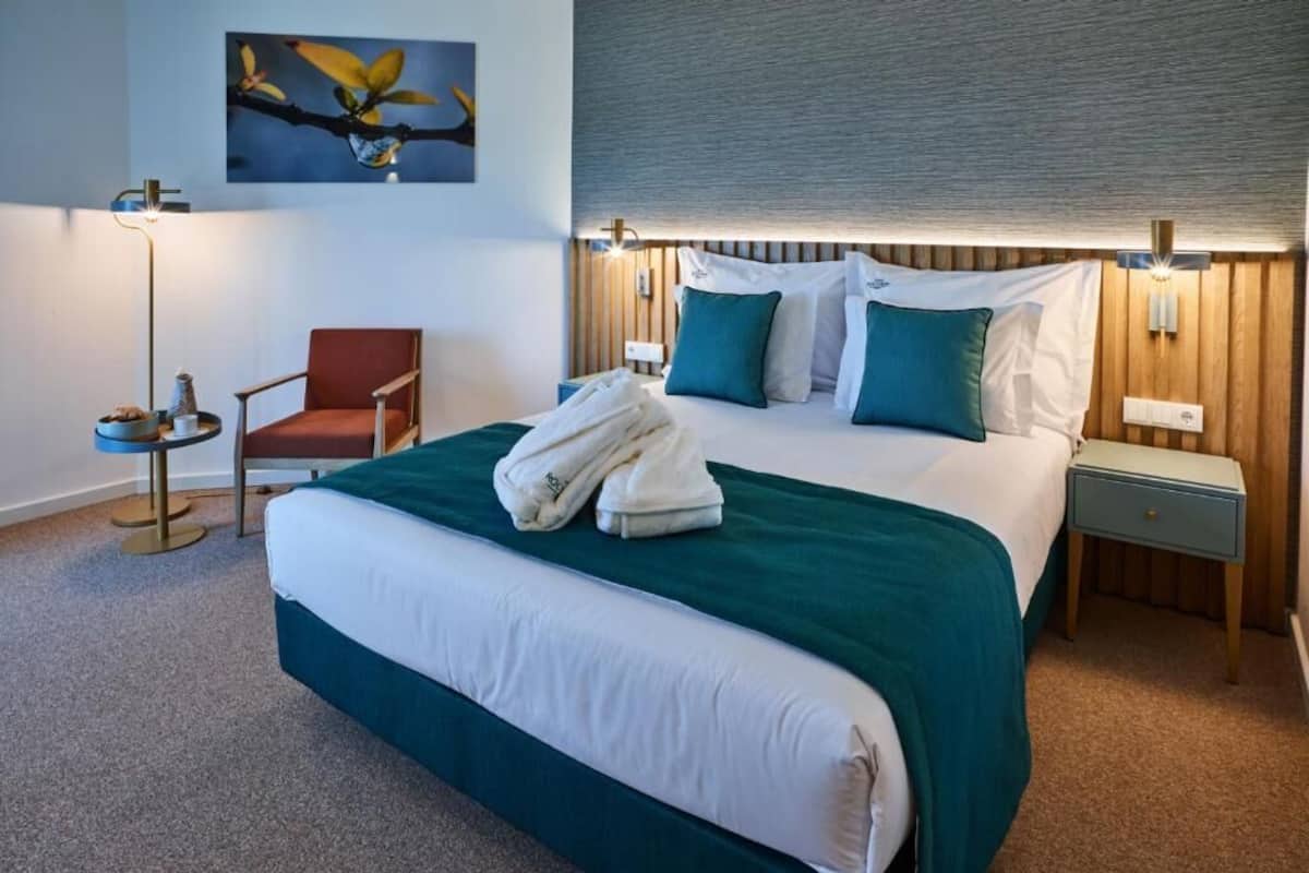 A hotel room with a large bed, a chair, a bedside table, a lamp, and a wall-mounted artwork.