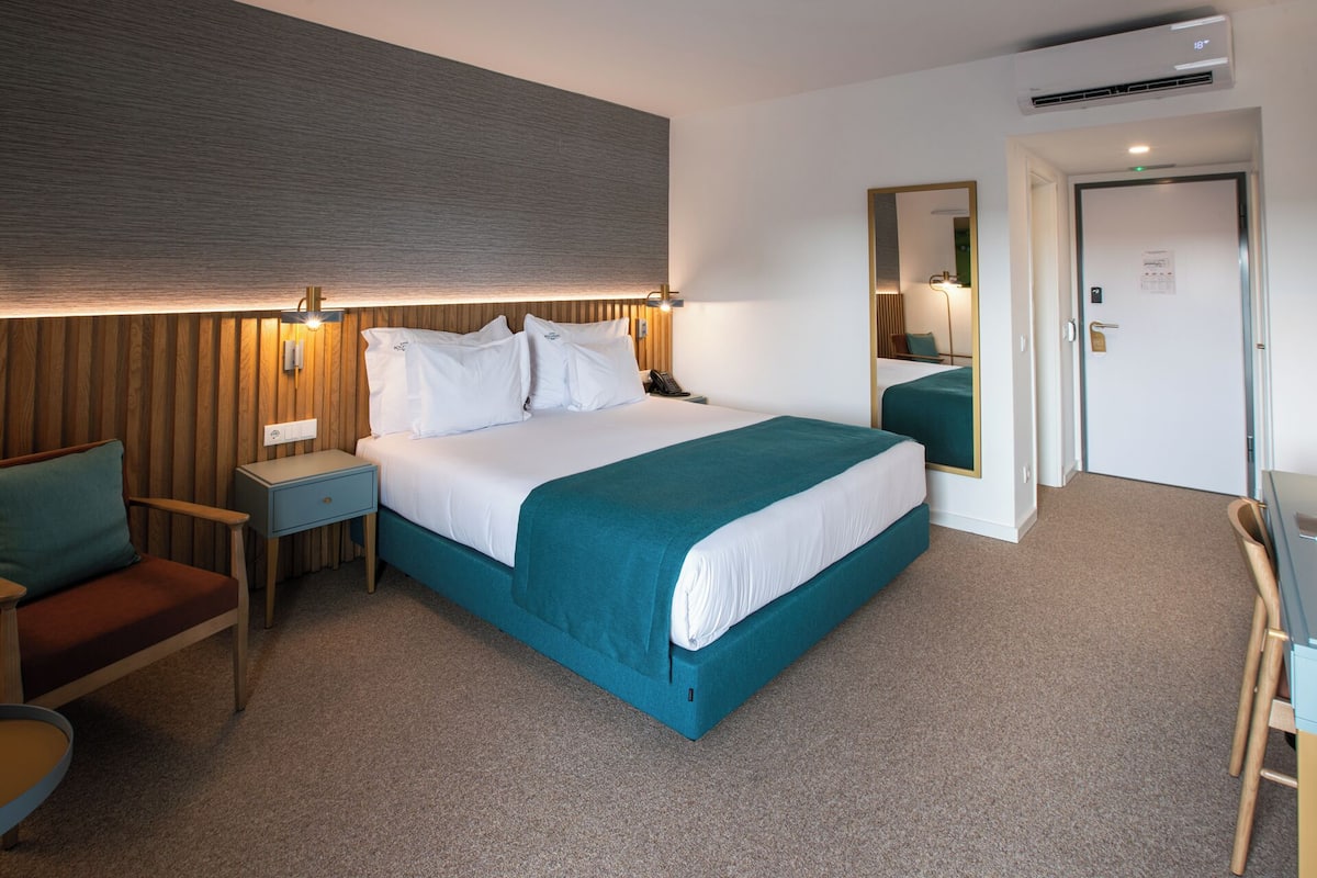 A hotel room with a large bed, a desk, a chair, a mirror, and a door.