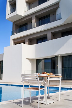 A modern building with a pool area featuring white chairs and a table with breakfast items.