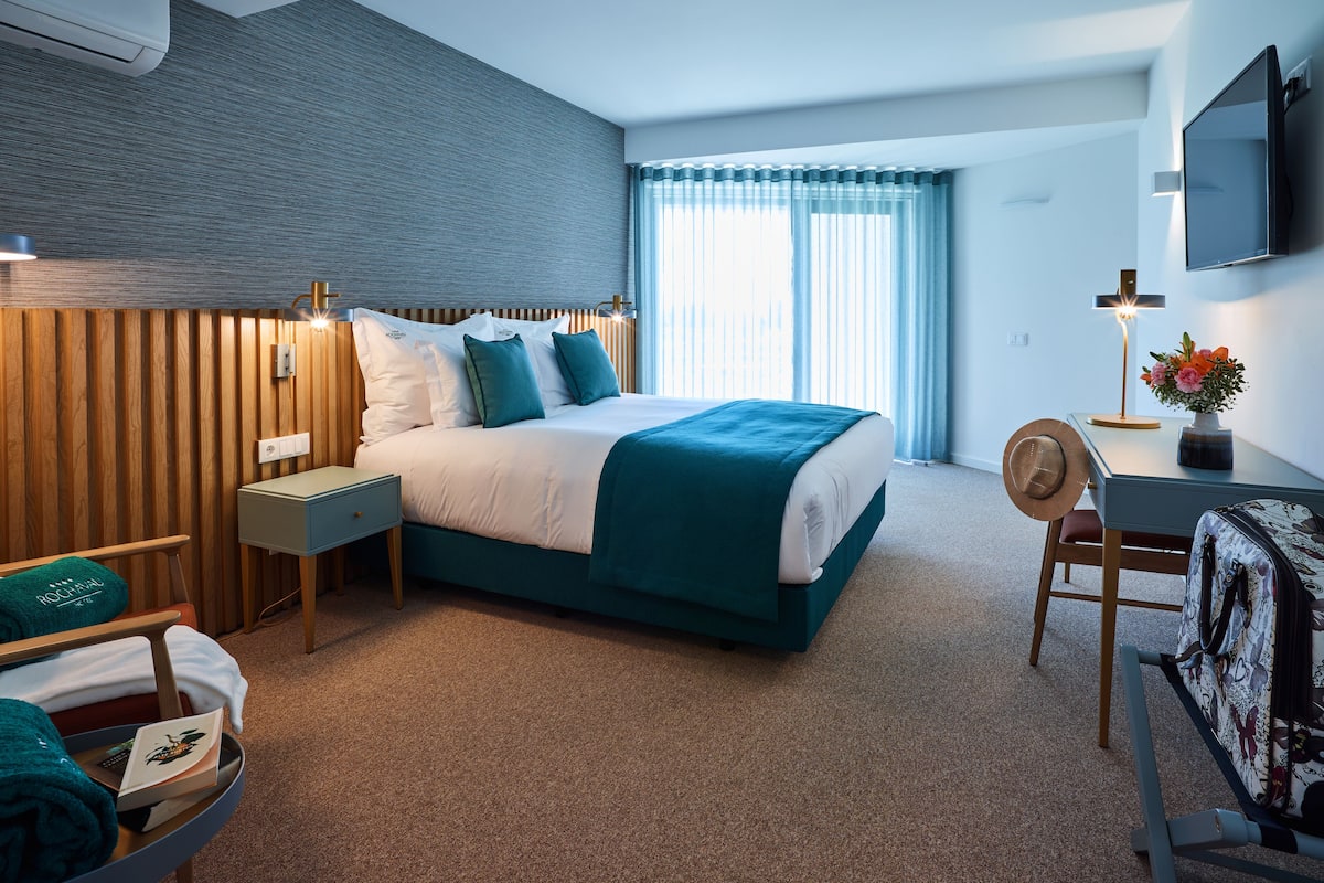 A hotel room with a large bed, a desk, a chair, a TV, and a window with curtains.