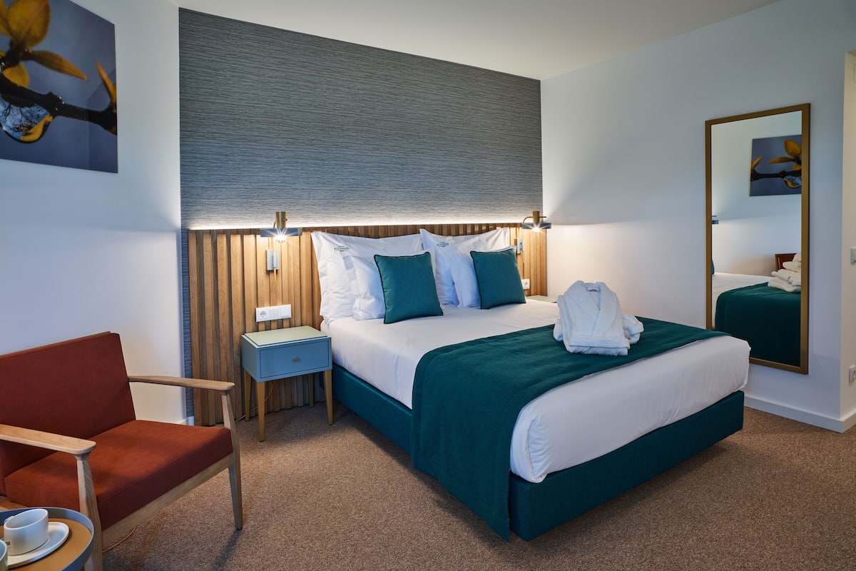 A modern hotel room with a neatly made bed, a wooden nightstand, a red armchair, and a wall-mounted mirror.