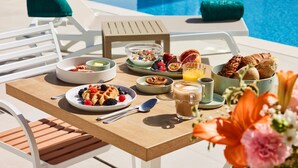 A breakfast table with various dishes, a pool in the background, and a chair with a towel. - Rochavau Hotel (Portimão)
