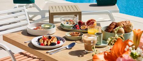 A breakfast table with various dishes, a pool in the background, and a chair with a towel.