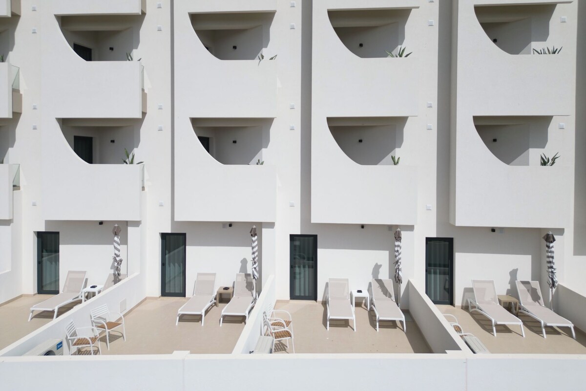 A modern building with multiple balconies, each with a small window and a plant. There are several lounge chairs and tables on the ground level.