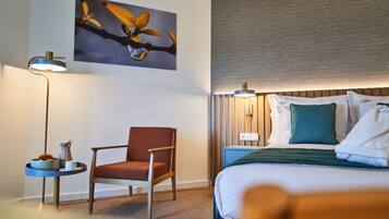 Hotellihuone, jossa on sänky, tuoli, yöpöytä, lamppu ja taulu seinällä.