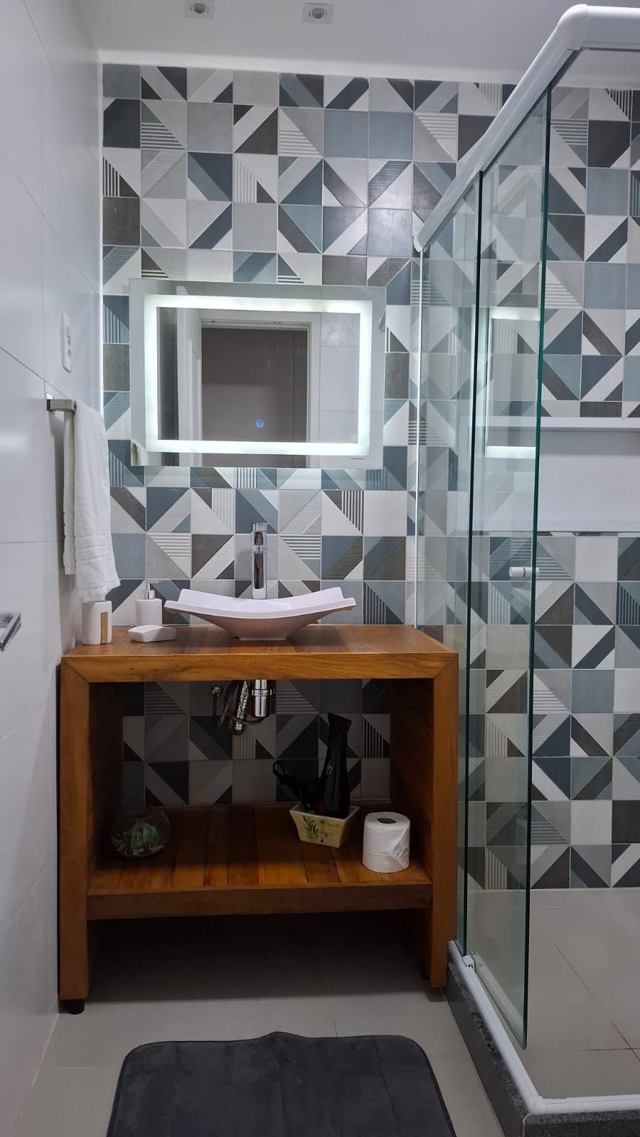 Une salle de bain moderne avec un mur recouvert de carreaux aux motifs géométriques, un meuble-lavabo en bois avec un miroir et une cabine de douche en verre.