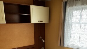 A single bed with white bedding, a bedside lamp, a small table, and a cabinet above the bed. - Mobil Home Gère Belesten (Gère-Bélesten)