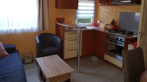A compact kitchen with a gas stove, oven, and a small dining table. There is a single chair and a sofa in the living area. - Mobil Home Gère Belesten (Gère-Bélesten)