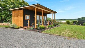 A modern wooden cabin with a covered porch, a gravel driveway, and a view of a lake.