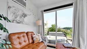 A modern living room with a brown leather sofa, a wooden coffee table, and a large window offering a view of a hot tub and greenery.