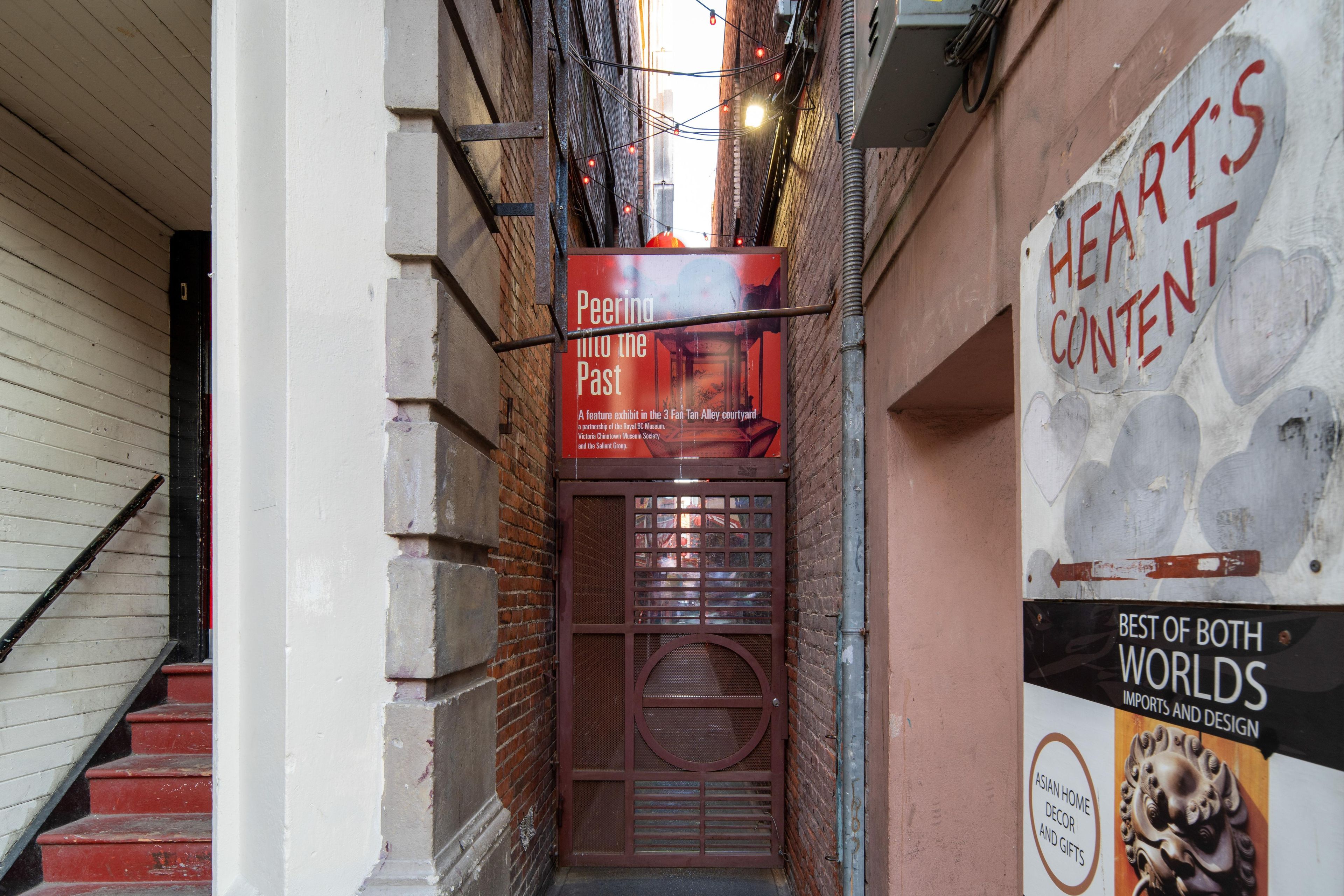 A narrow alleyway with a red sign for 'Hearts Content' and a poster for 'Best of Both Worlds Imports and Design.'