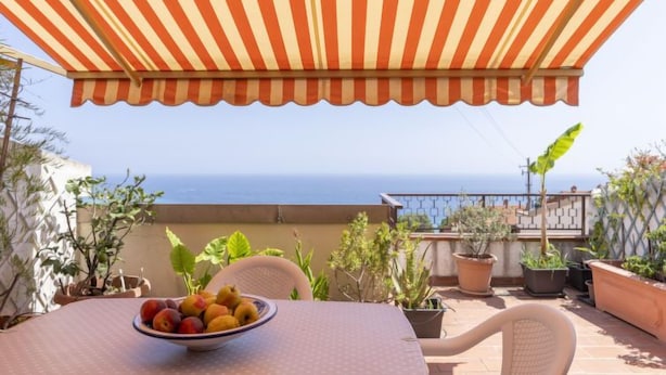 Eine Terrasse mit Tisch unter einem gestreiften Sonnensegel, einer Obstschale und Topfpflanzen.