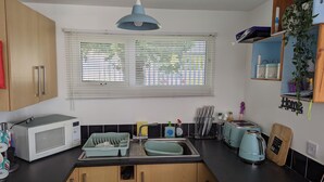 A compact kitchen with a microwave, toaster, and sink. There is a window with blinds, a hanging light fixture, and a shelf with kitchen utensils.