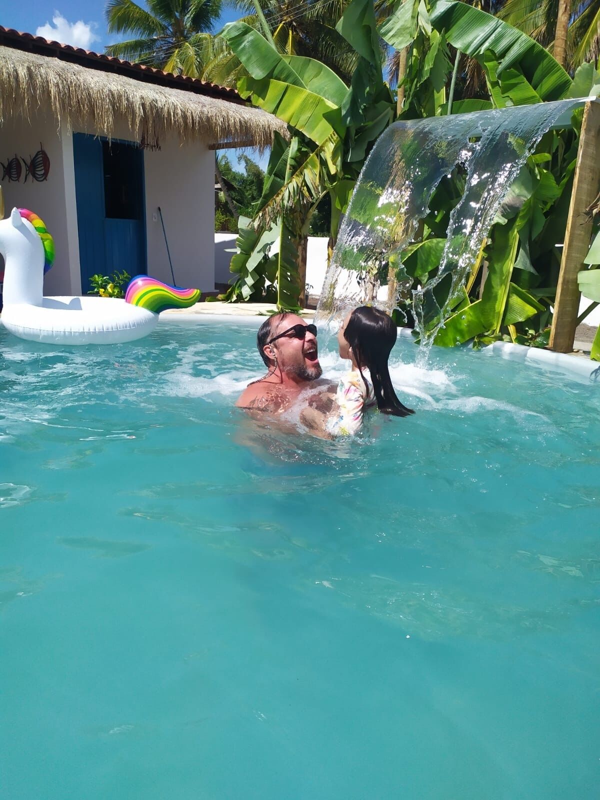 Um homem em uma piscina, com um cão, cercado por plantas tropicais e uma construção com telhado de palha.