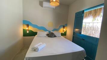 Una cama bien hecha con una toalla enrollada, un mural en la pared con un sol y olas, y una puerta azul.