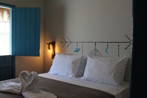 Un lit avec des oreillers blancs et une serviette, un mur décoré d’un croissant de lune et d’une étoile, et une fenêtre avec des volets bleus.