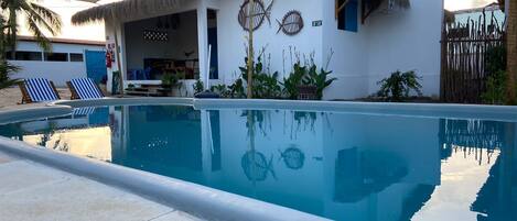 A poolside area with a clear blue pool, a thatched-roof building, and two lounge chairs.