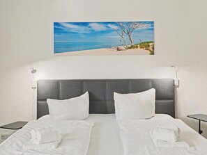 Camera d'albergo con un letto, due cuscini, un comodino e una lampada a parete, oltre a un quadro raffigurante una spiaggia.