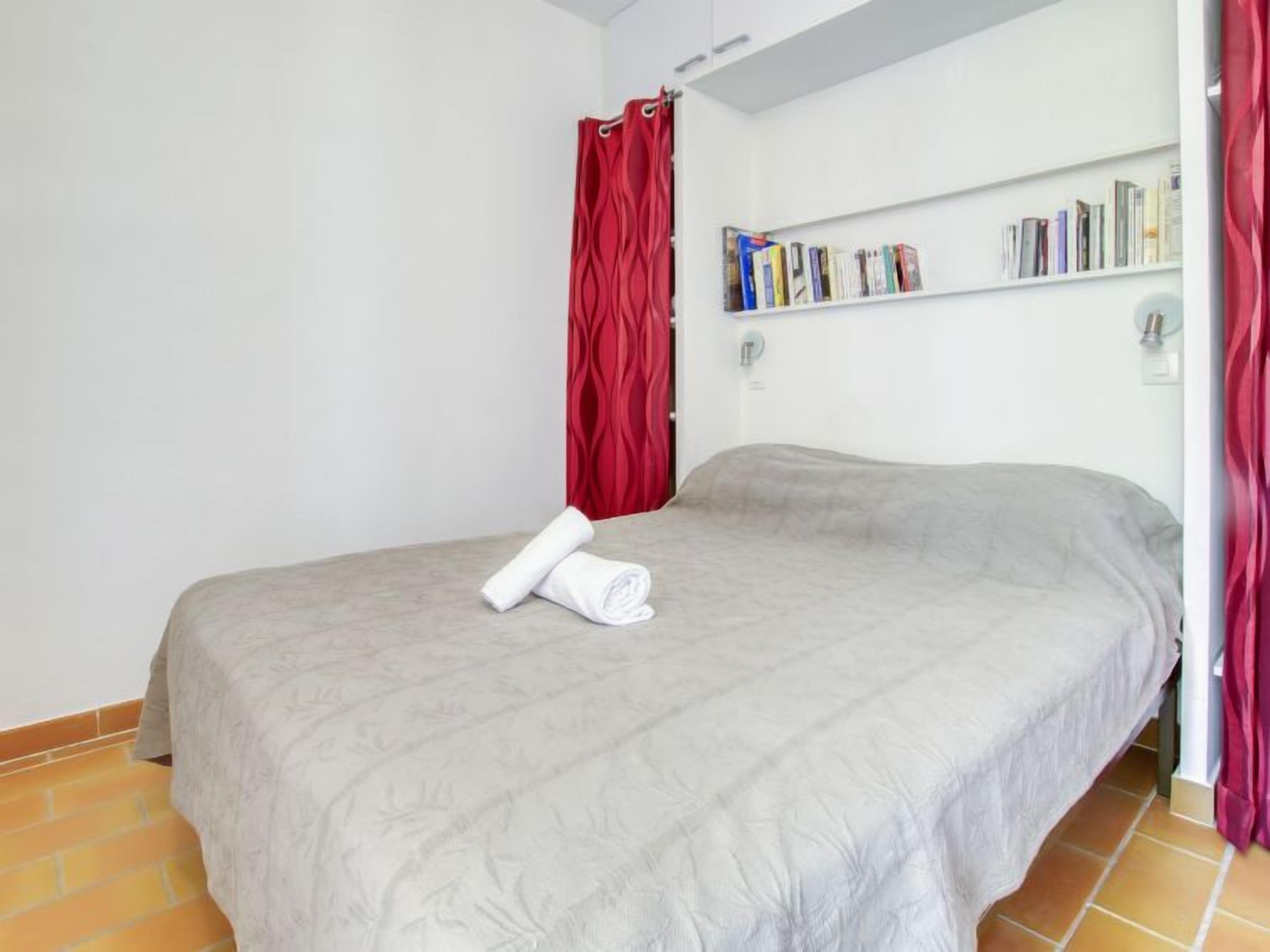 Een opgemaakt bed met een grijze deken en een opgerolde witte handdoek, een boekenplank met boeken, en een rood gordijn.