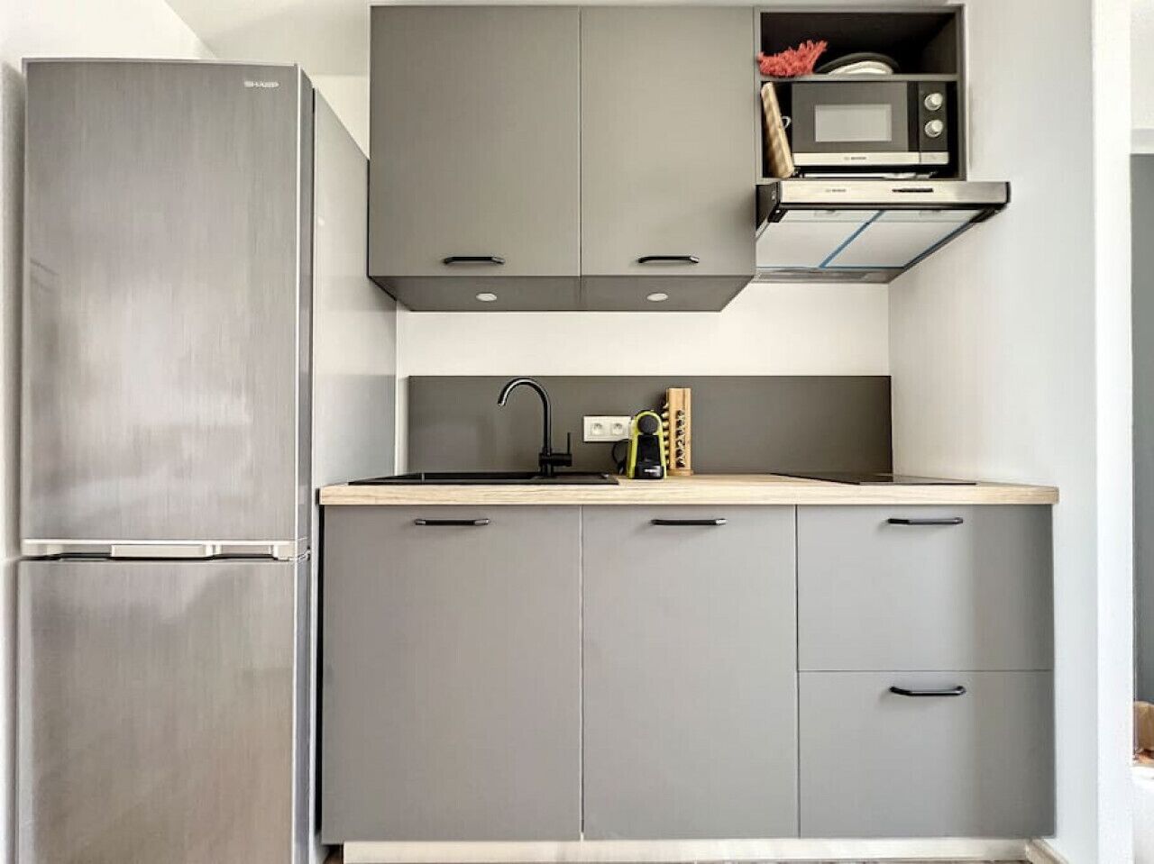 Cocina compacta con refrigerador de acero inoxidable, gabinetes grises, microondas y horno empotrado.
