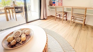 A room with a large window, wicker furniture, a round table with decorative items, and a beach-themed painting. - Hello Summer! beachfront studio in the heart of Orient Bay, pool direct access (Orient Bay)