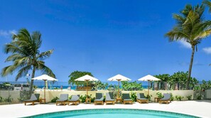 A poolside area with lounge chairs and umbrellas, a clear blue sky, and palm trees. - Hello Summer! beachfront studio in the heart of Orient Bay, pool direct access (Orient Bay)