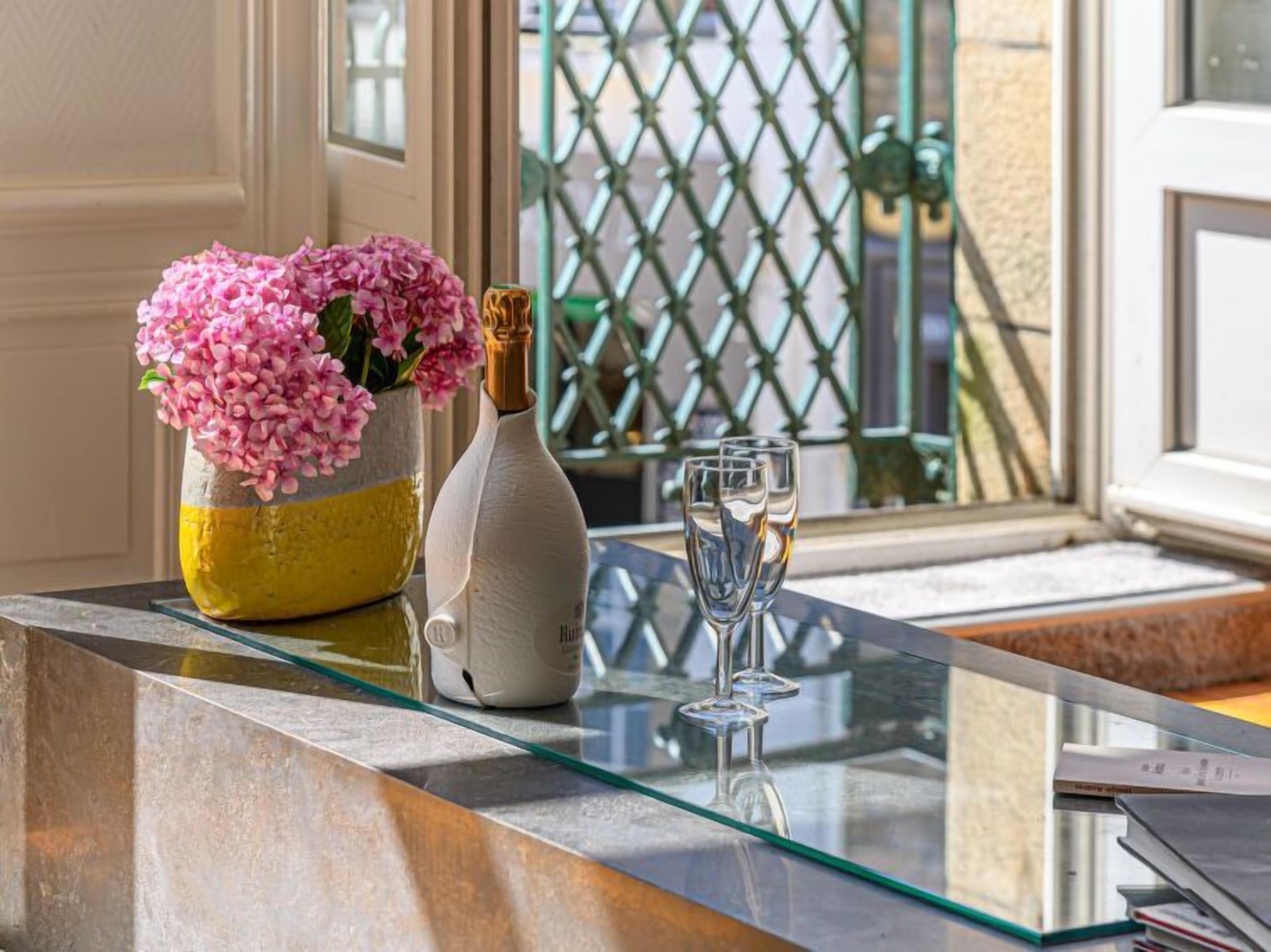 A glass-topped table with a vase of pink flowers, a bottle, and two glasses.