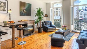 Un saló modern amb sòl de fusta, una zona de menjador amb taula i cadires, un sofà de cuir negre, un butac armchair negre, una planta en test i un quadre a la paret.