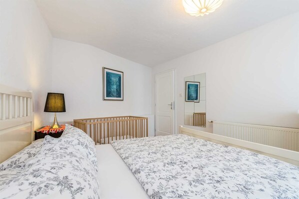 A bedroom with a crib, a bedside table with a lamp, a framed artwork, and a mirror.