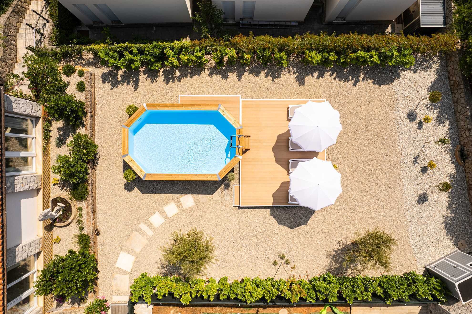 An aerial view of a rooftop pool area with a rectangular pool, two white umbrellas, and surrounding greenery.