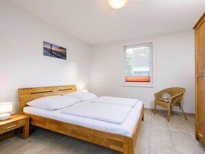 Ein Schlafzimmer mit einem hölzernen Bett, einem Nachttisch, einem Sessel, einem Fenster und einem Bild an der Wand.