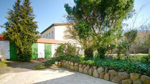 Un patio empedrado con un muro de piedra, un edificio blanco con puertas verdes y vegetación circundante.