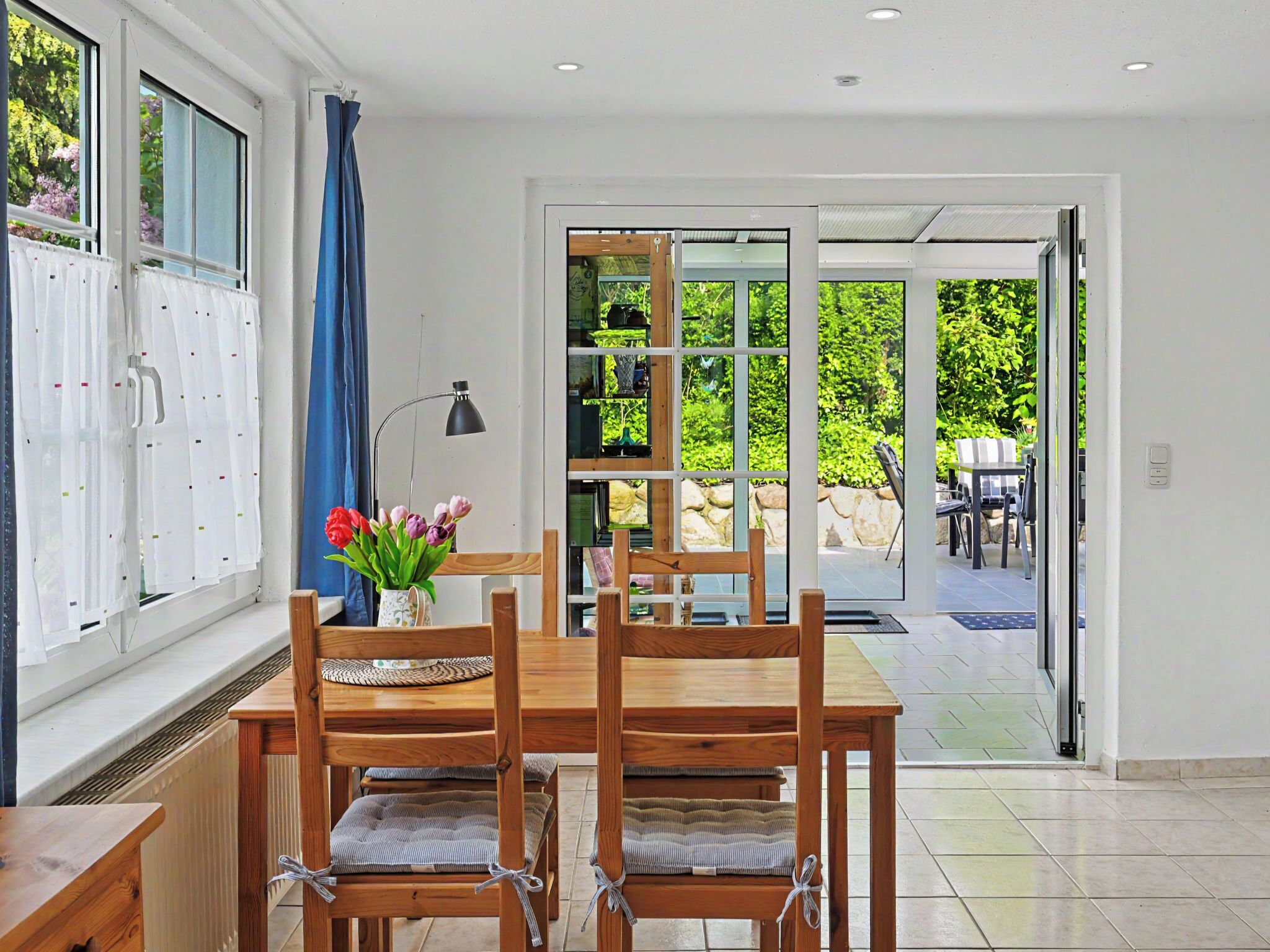 A dining area with a wooden table and chairs, a vase with flowers, and a view of an outdoor patio with a table and chairs.