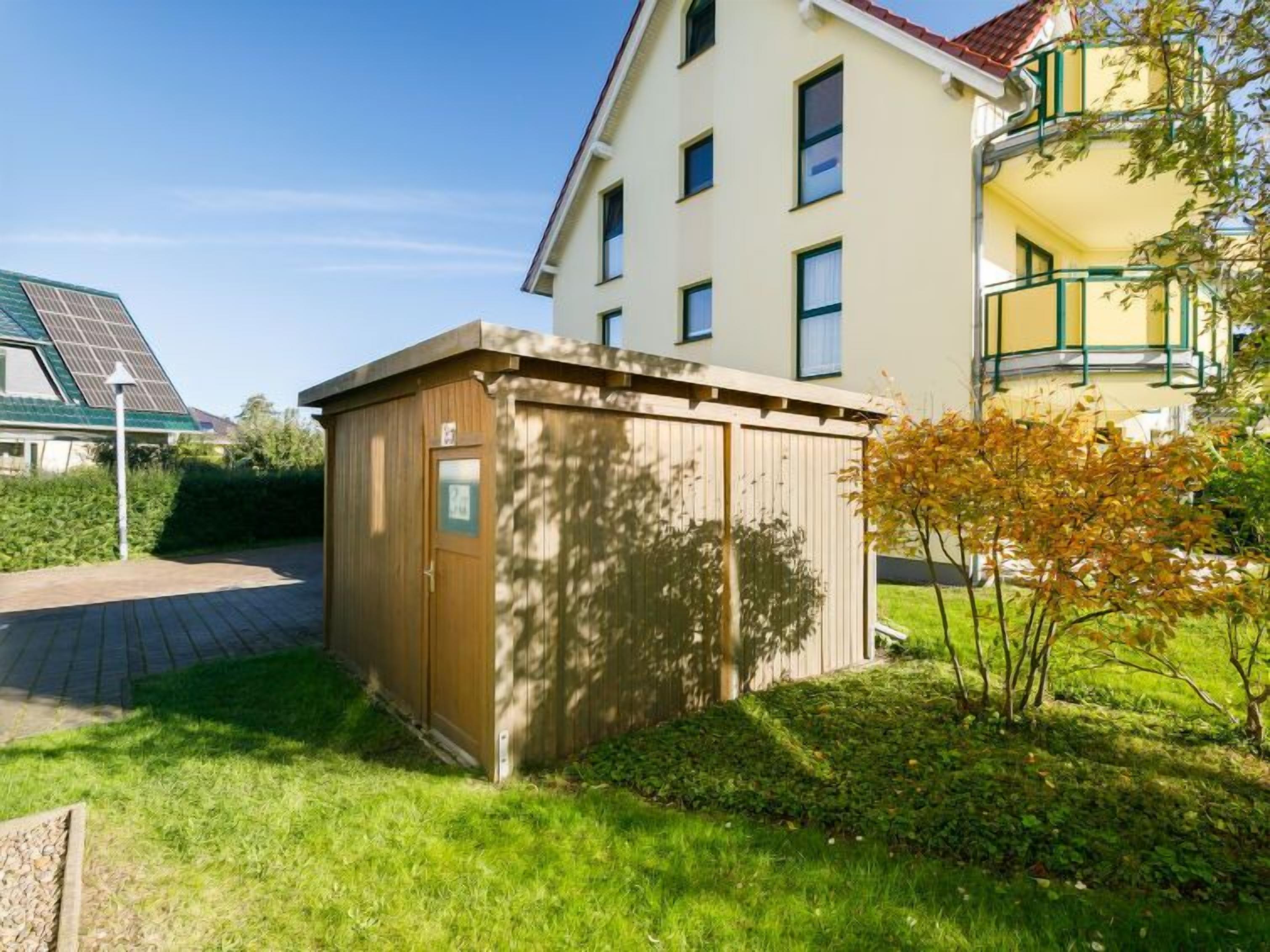 Eine kleine Holzhütte in einer grünen Wiese, im Hintergrund ein Gebäude.