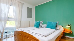 Ein Schlafzimmer mit einem hölzernen Bett, weißer Bettwäsche, türkisfarbenen Kissen, einer grünen Wand, einem Fenster mit transparenten Vorhängen und einer Nachttischlampe.