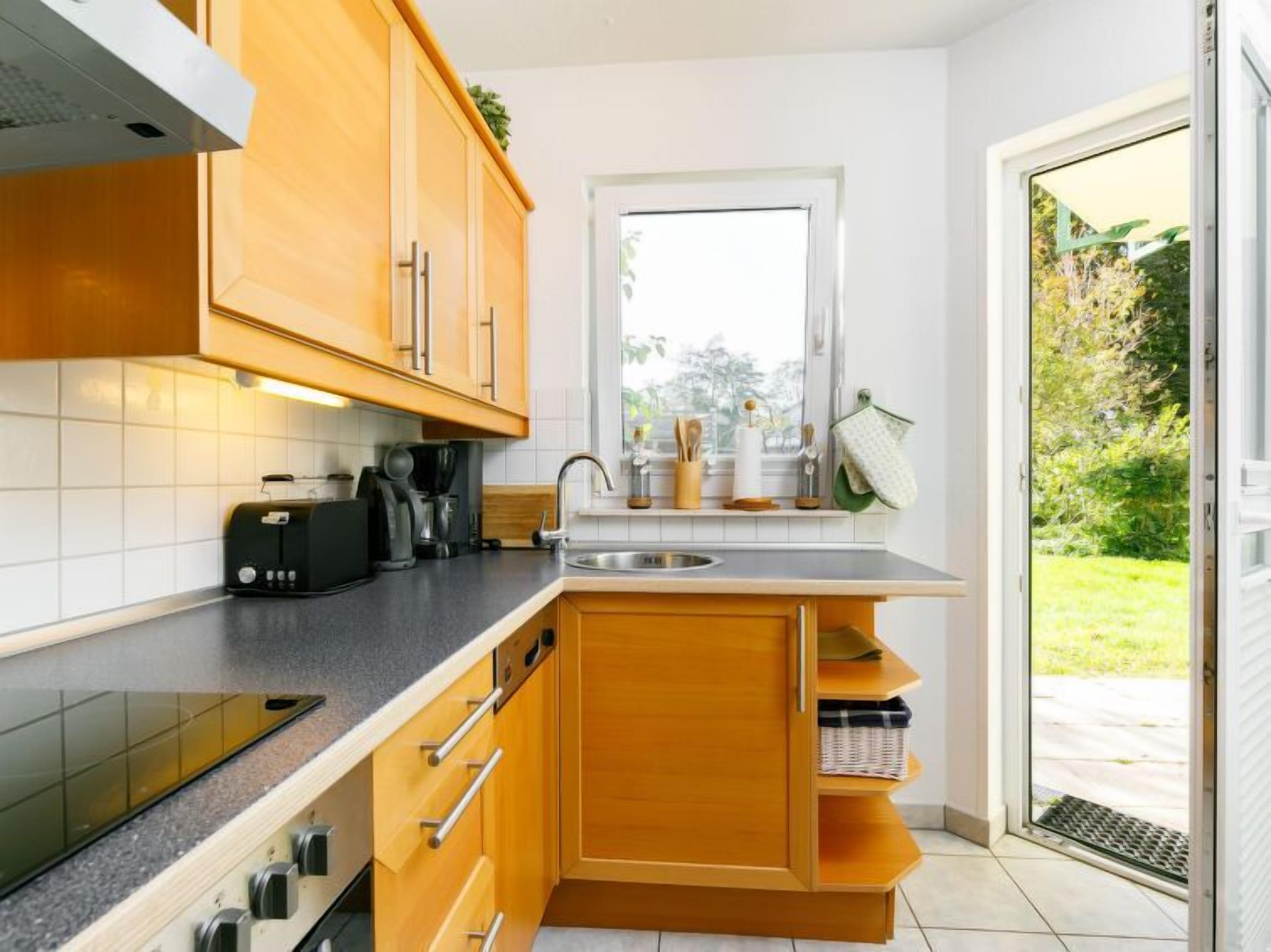 A compact kitchen with wooden cabinets, a built-in oven, a coffee maker, and a window overlooking a garden.