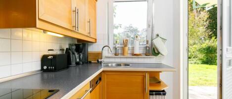 A compact kitchen with wooden cabinets, a built-in oven, a coffee maker, and a window overlooking a garden.