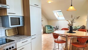 A modern kitchen with a microwave, stove, and wooden cutting board. There is a dining area with a round table and orange chairs. A green armchair and a vase with flowers are also visible. - Zaunkönig by Interhome (Zinnowitz)
