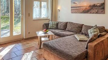 Ein gemütliches Wohnzimmer mit einer Eckcouch, einem kleinen Tisch mit Obstschale und einem Gemälde, das eine Strandansicht zeigt.