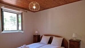 Een tweepersoonsbed met witte lakens en handdoeken, een houten hoofdbord, twee nachtkastjes met lampen, een houten plafond en een raam met uitzicht op groen.