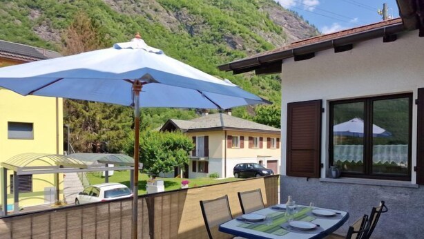 Een terras buiten met een tafel, stoelen, een blauwe parasol en uitzicht op een berg en huizen op de achtergrond.