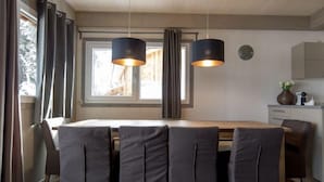 A dining area with a wooden table and four grey upholstered chairs. There are two black pendant lights hanging above the table. A window with curtains is visible behind the table, and a clock is mounted on the wall to the right.