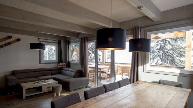 A modern living room with a wooden dining table, a sofa, and a large window with a view of trees.