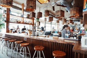 A bar with wooden stools, hanging wicker baskets, and a counter with various bottles and a cash register.