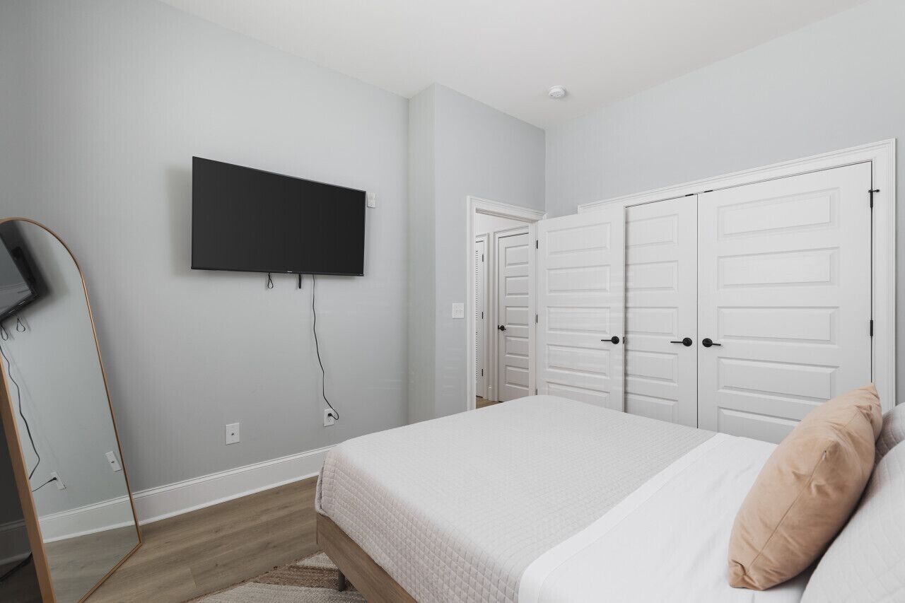 Un dormitorio con cama, televisor montado en la pared y un conjunto de puertas blancas.