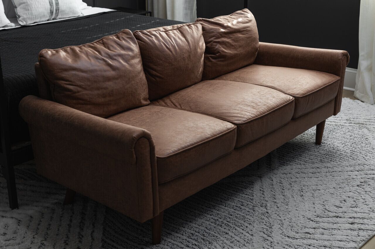 A brown leather sofa with wooden legs in a room with a dark wall and a white rug.