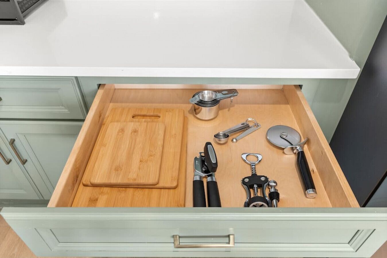 A kitchen drawer with a wooden cutting board, a metal measuring cup, a can opener, a bottle opener, and a pair of scissors.